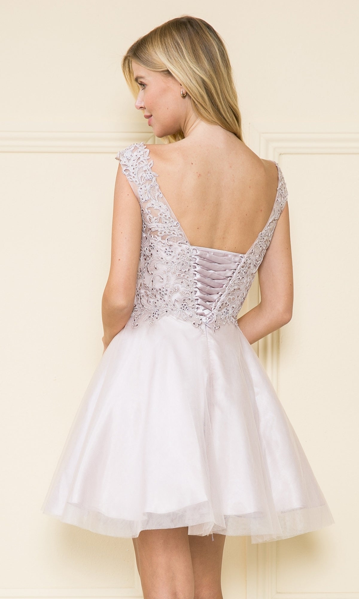 A woman wearing the Lace-Bodice Short Babydoll Homecoming Dress 8902, featuring a fitted bodice and lace-up back, stands facing slightly away from the camera.